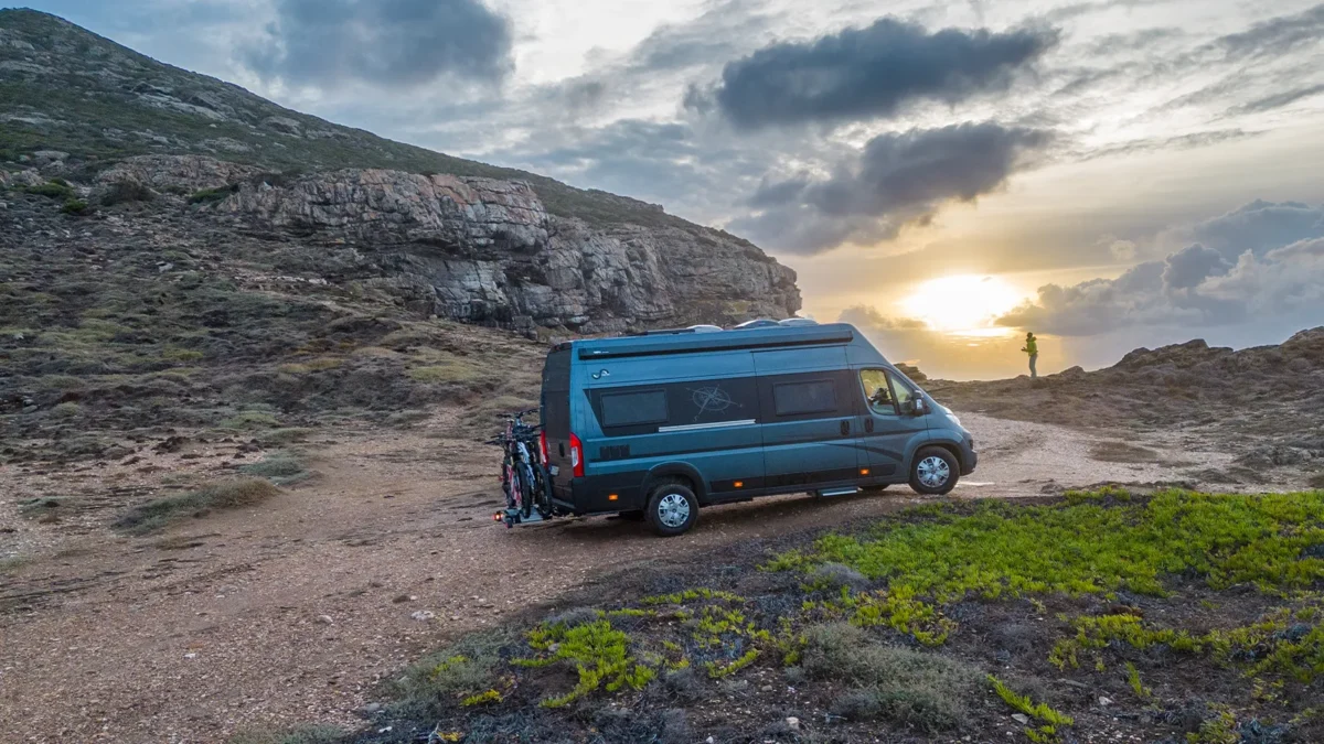 Blauer Camper steht auf einer Gebirgsstraße bei Sonnenuntergang.