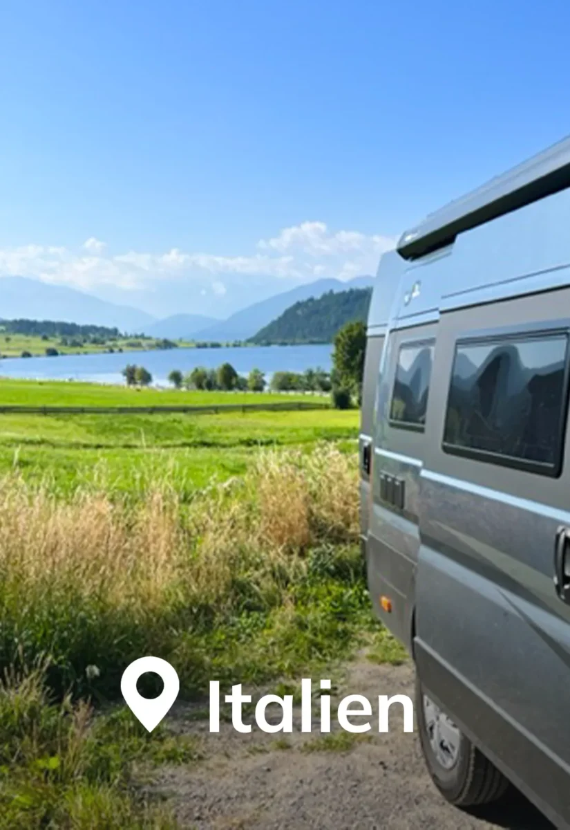 Seitenwand eines blauen Campers mit Blick auf eine Wiese und einen Bergsee.