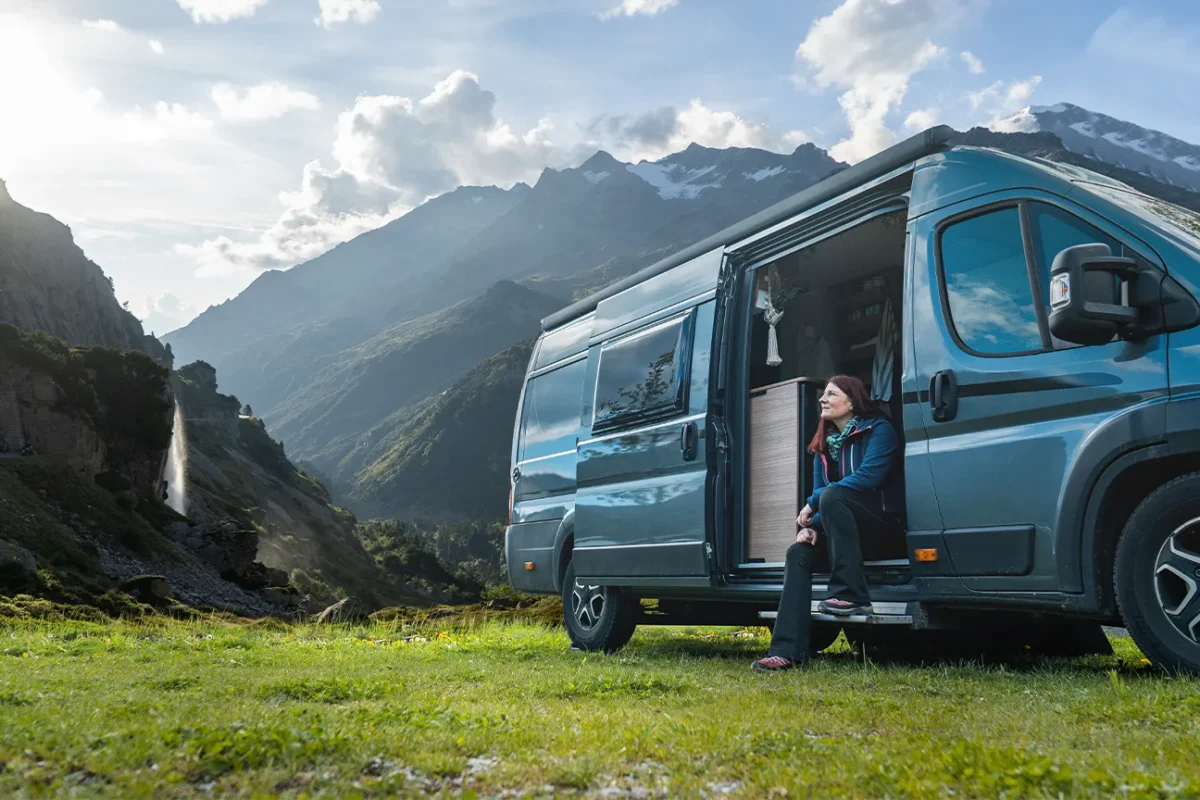 Frau sitzt in geöffneter Tür eines beim Autohaus Hempel gemieteten Campers in einer Berglandschaft.