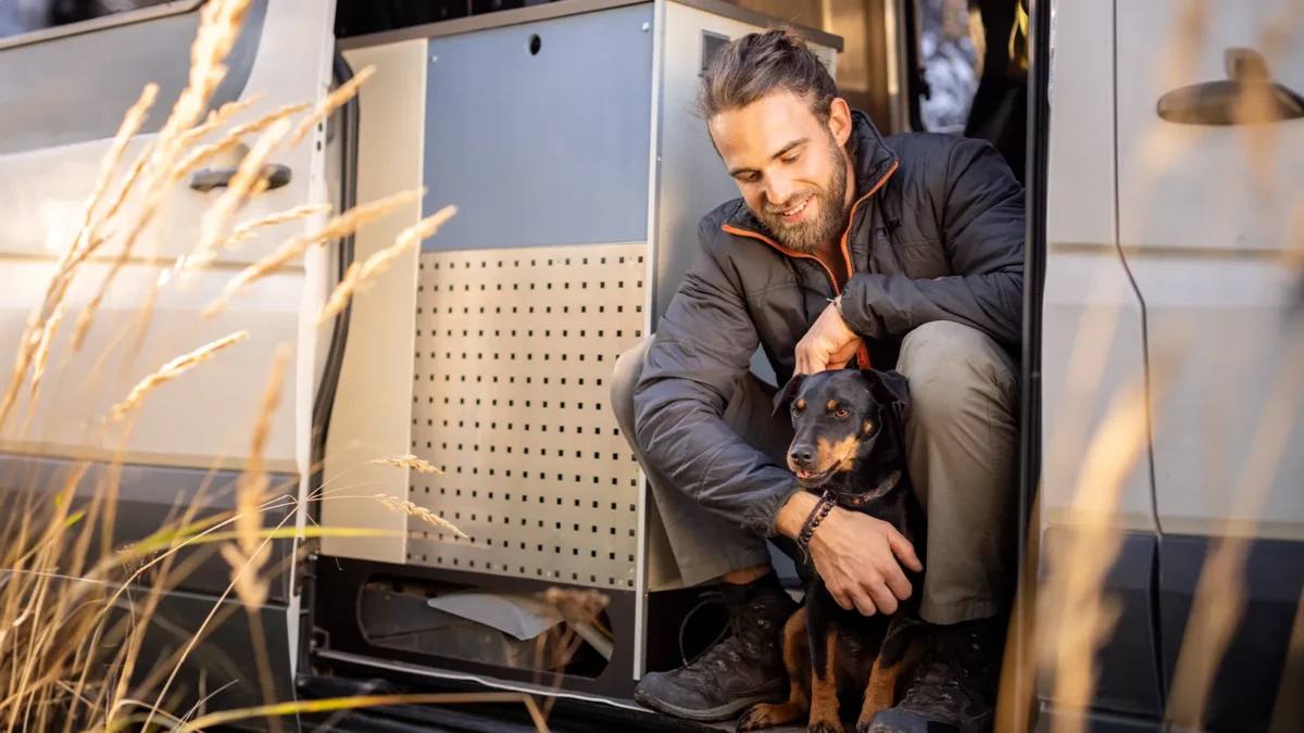 Mann sitzt mit einem schwarzen Hund im Arm in der geöffneten Seitentür eines silbernen Campervans.