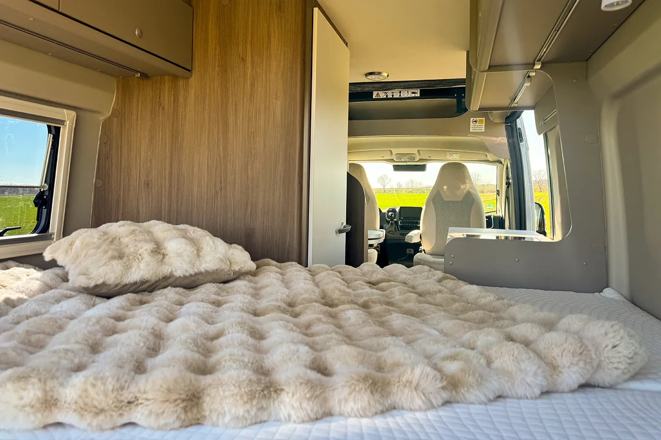 Komfortables Camper-Bett mit Kissen und Blick auf ein grünes Feld durch die geöffnete Hecktür des Fiat Carado Campers