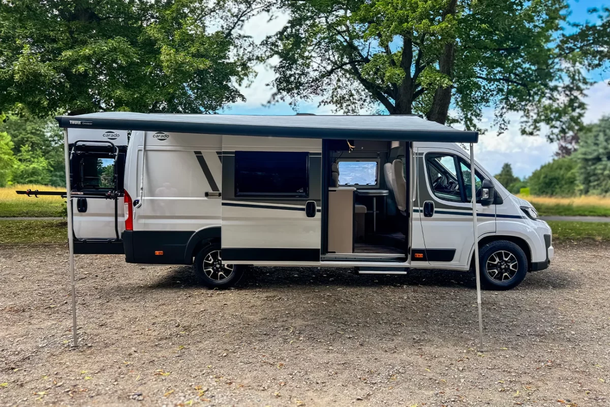 Seitliche Sicht auf einen weißen Fiat Carado Typ 640 Camper mit ausgefahrener Markise, der auf einer grünen Wiese steht.