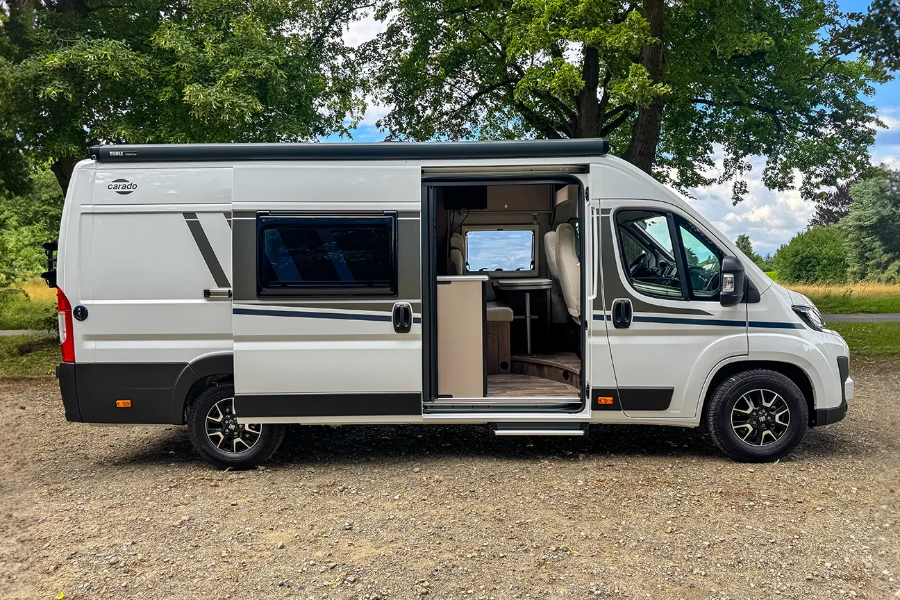Schräge Sicht auf einen weißen Fiat Carado Typ 640 Camper, der mit geöffneter Seitentür auf einer grünen Wiese steht.