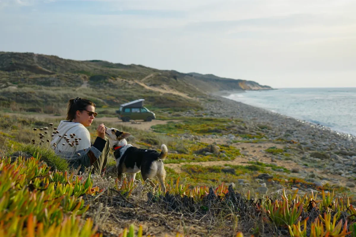 Frau sitzt mit ihrem Hund im Gras an einer grünbewachsenen Küste mit einem Campervan im Hintergrund.